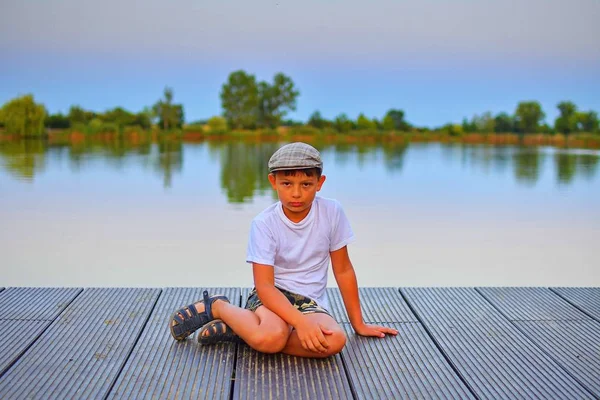 桟橋に座っている小さな男の子 木製の桟橋に座っている小学校低学年少年 夏と子供の頃のコンセプトです 湖でベンチの子 休暇の夕方の間に黄金の時間 — ストック写真