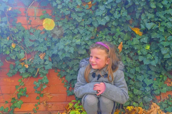 Kis lány visel retro kabát és a guggolás előtt tégla fal, ősszel. Borostyán fal ősszel. Őszi koncepció. Lens flare — Stock Fotó