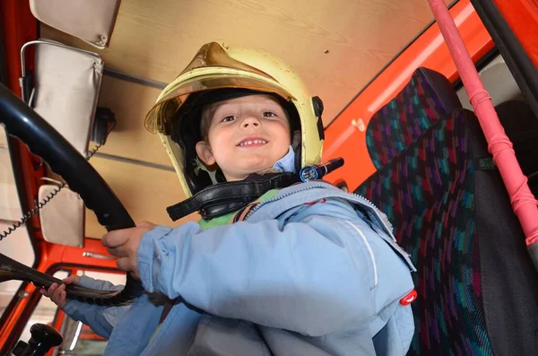 Mały chłopiec zachowujący się jak strażak. Chłopak siedzi w prawdziwym wozie strażackim. Szczęśliwy uroczy chłopiec ze strażackim kapeluszem siedzący w czerwonym wozie strażackim. Marzy o przyszłej profesji. Bezpieczeństwo pożarowe, życie — Zdjęcie stockowe