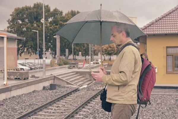 Portré egy érett utazóról, aki okostelefonon SMS-ezik egy esernyővel a pályaudvar peronján. Befejezetlen vasútvonal. Amikor a vonat megérkezik... A hiábavaló helyzet vicces fogalma — Stock Fotó