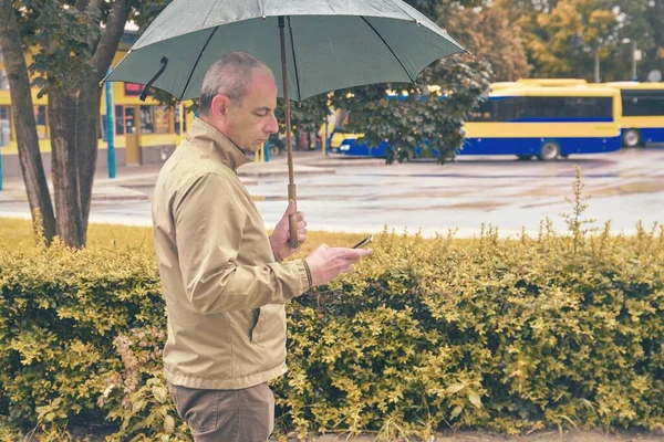 A man standing at the bus station with an umbrella waiting for a bus. Man waiting on a bus, texting on mobile phone in a rainy day. Rainy day in a local town