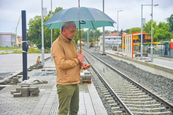 Retrato de um homem maduro com um guarda-chuva em pé na plataforma de uma estação de trem. Linha ferroviária inacabada. Quando o comboio chegar... Conceito engraçado de situação sem esperança — Fotografia de Stock