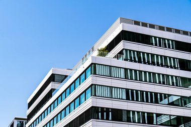 Çağdaş ofis binası. Yansıtıcı mavi pencereleri, beyaz perisi ve parlak gökyüzü altında minimum sarı aksanları var..