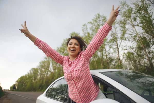 Cute laughing young woman hanging out her head from a car through the open window enjoying the freedom of the breeze in her hair as it travels along a rural road — Stock Photo, Image