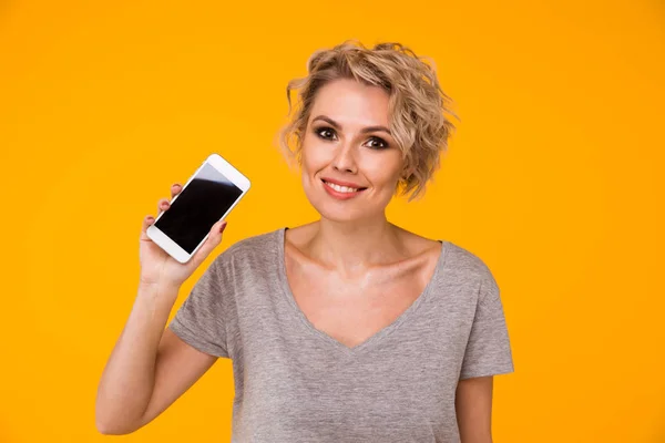 Šťastné blond žena v svetr zobrazení prázdné smartphone obrazovky a ukazoval na něj při pohledu na fotoaparát s otevřenými ústy nad žlutá — Stock fotografie