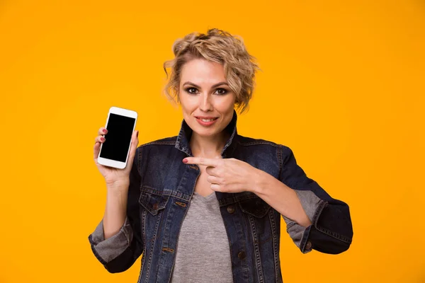 행복 한 금발 여자 스웨터 빈 스마트폰 화면을 표시 하 고 노란색 배경 위에 오픈 입으로 카메라를 보고 하는 동안 그것에 가리키는 — 스톡 사진