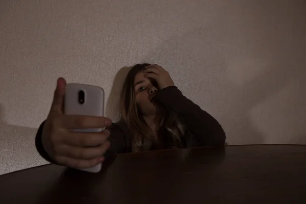 Único adolescente triste segurando um telefone celular lamentando sentado na cama em seu quarto com uma luz escura — Fotografia de Stock