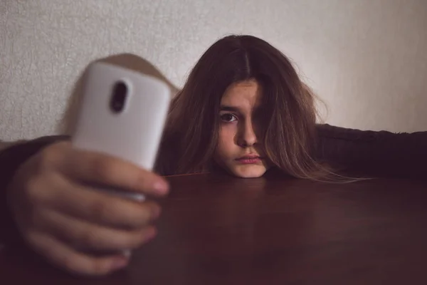 Soltera adolescente triste sosteniendo un teléfono móvil lamentándose sentada en la cama en su dormitorio con una luz oscura — Foto de Stock