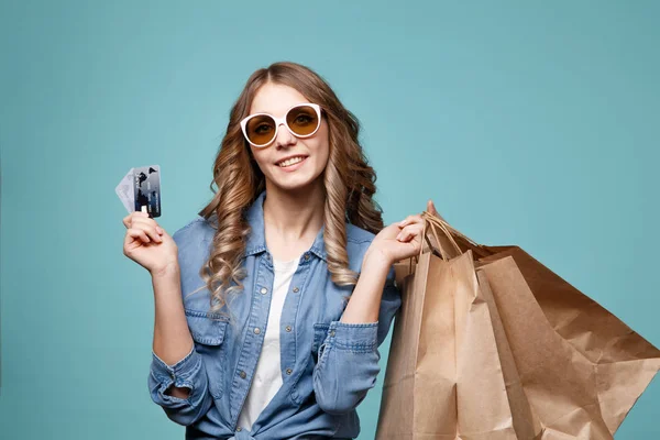 Portrét veselé krásná dívka, která nosí šaty a sluneční brýle drží nákupní tašky a ukazuje kreditní kartu izolované nad modrým pozadím — Stock fotografie
