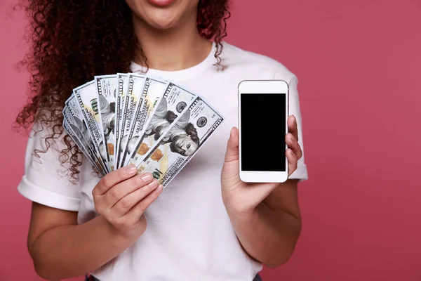 Portret podekscytowany młode kobiety afrykańskie trzymając kilka banknotów pieniądze i patrząc na telefon komórkowy na białym tle różowy — Zdjęcie stockowe