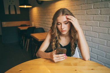 Arayan bir kafede oturan üzgün genç kız yorgun onun telefon, siyah telefonunu izlerken mutsuz kız,