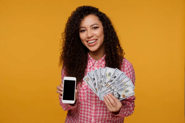Portret podekscytowany młode kobiety afrykańskie trzymając kilka banknotów pieniądze i patrząc na telefon komórkowy na białym tle nad różowy — Zdjęcie stockowe