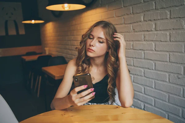 Triste giovane ragazza seduta in un caffè cercando stanco il suo telefono, ragazza infelice guardando il suo telefono nero , — Foto Stock