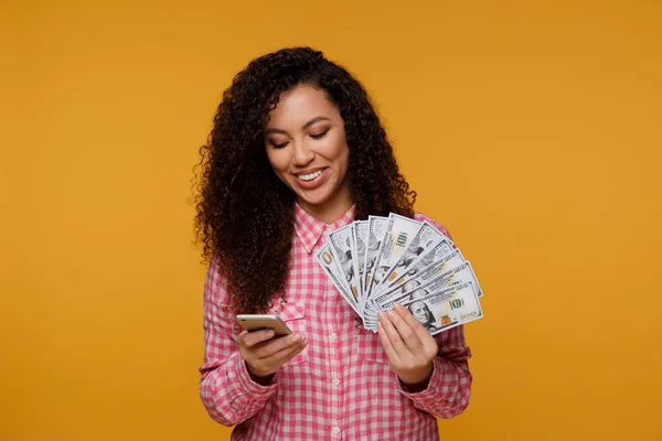 Young lady isolated over blue background. Looking camera showing display of mobile phone holding money — Stock Photo, Image