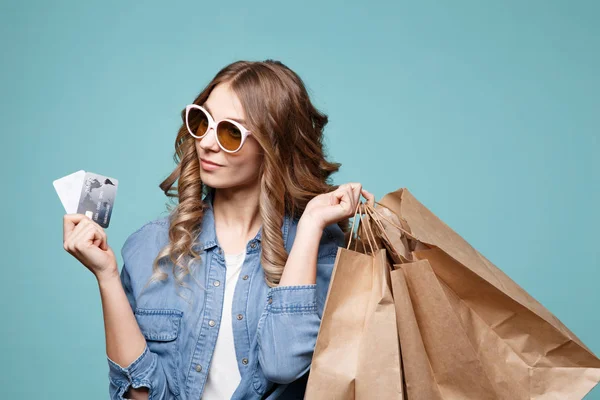 Πορτρέτο του ένα χαρωπό όμορφο κορίτσι που φοράει φόρεμα και γυαλιά ηλίου, κρατώντας τσάντες για ψώνια και δείχνει την πιστωτική κάρτα που απομονώνονται σε μπλε φόντο — Φωτογραφία Αρχείου