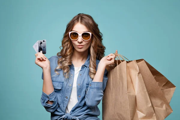 Portret van een vrolijke mooi meisje dragen jurk en zonnebril boodschappentassen houden en tonen creditcard geïsoleerd op blauwe achtergrond — Stockfoto