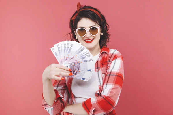 Portret uśmiechniętej dziewczyny, trzymając kilka banknotów pieniędzy i patrząc na telefon komórkowy na białym tle różowy — Zdjęcie stockowe