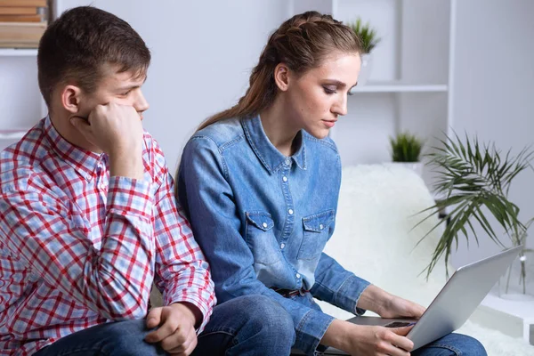 Problem in couple. Upset man sitting on a sofa near his woman watching laptop.