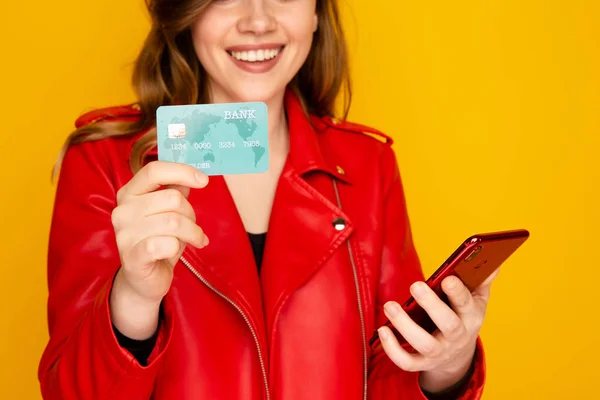 Nő a piros kabát kezében hitelkártya és a telefon izolált a sárga háttér. — Stock Fotó