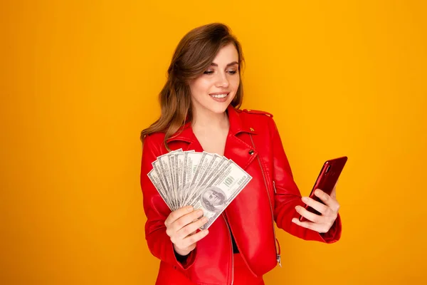Dość uśmiechnięta kobieta w czerwonej kurtce trzymając pieniądze i telefon na żółtym tle. — Zdjęcie stockowe