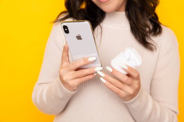 Mutlu esmer kadın telefon ve airpods tutan sarı stüdyo izole.