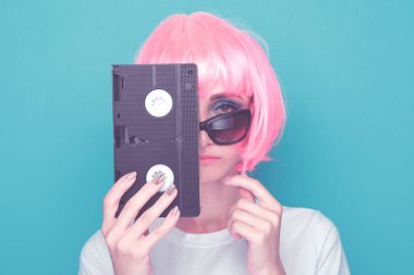 90'ların stil konsepti. Mavi renkli stüdyoda duran gözlük ve kaset tutan pembe dolu ile genç kadın.