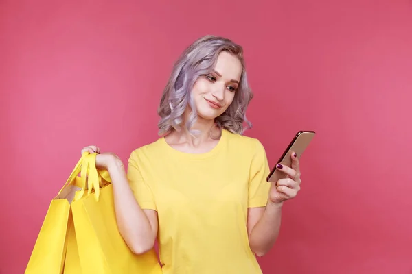 Online winkelen concept. Schattige jonge vrouw kijken naar haar telefoon en het houden van tassen geïsoleerd. — Stockfoto