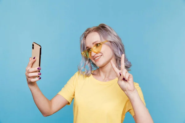 Vtipné dívka ve žlutém oblečení a brýle dělat vlastní foto na svém telefonu v modrém pokoji. — Stock fotografie