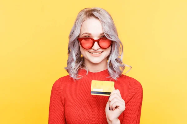 Blond woman in red glasses holding credit card isolated in the yellow studio. — Stockfoto