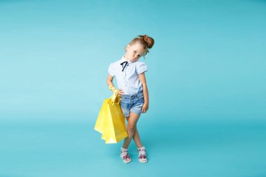 Mavi stüdyoda büyük, parlak, sarı kağıt torbaları tutan sevimli ve komik bir kız..