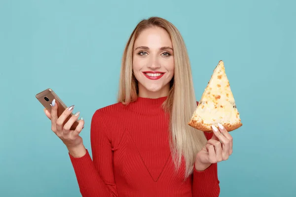 Female person eating pizza and using phone isolated over the blue background.