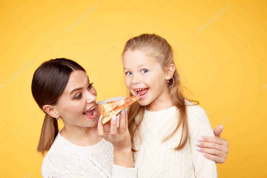 Mother and daughter eating pizza for lunch isolated over the yellow background.
