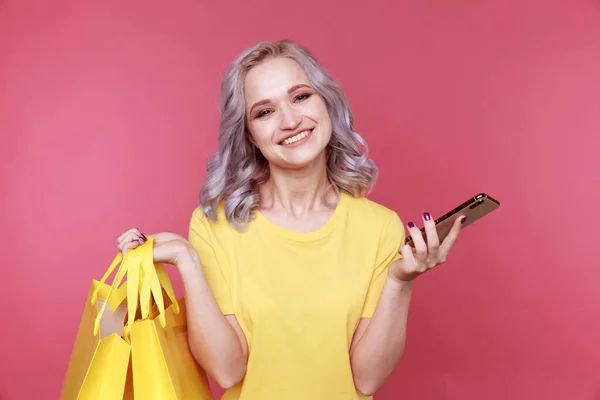 Jonge stijlvolle vrouw in de gele t-shurt holding telefoon en dezelfde solor packege geïsoleerd. — Stockfoto