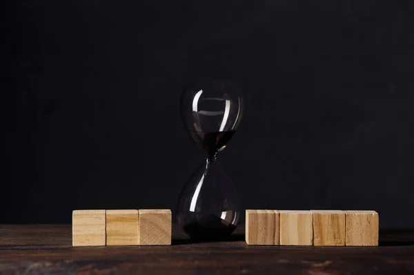 Reloj de arena o vidrio de arena entre bloques de madera cubo, lugar para el texto, en la mesa de madera con fondo oscuro —  Fotos de Stock