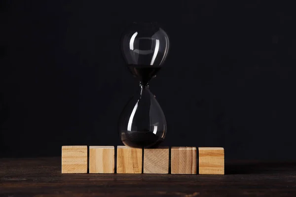Reloj de arena o vidrio de arena en bloques de madera cubo, lugar para el texto, en la mesa de madera con fondo oscuro —  Fotos de Stock