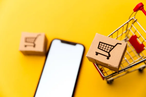 Vista superior sobre un carrito de la compra, cajas y teléfono móvil sobre un fondo amarillo. Concepto de compras en línea Smartphone — Foto de Stock