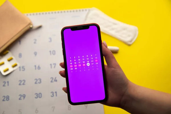 Concepto de ciclo menstrual. Calendario del mes con marcas y una aplicación móvil en la pantalla del smartphone. Anticonceptivos y toallas sanitarias en el fondo —  Fotos de Stock