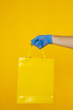 Tıbbi eldivenli kurye, sarı arka planda izole edilmiş alışveriş torbaları tutuyor..