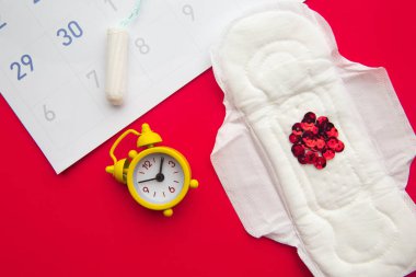 Hijyenik ped ve kırmızı zemin üzerinde sarı alarm saati olan adet takvimi.