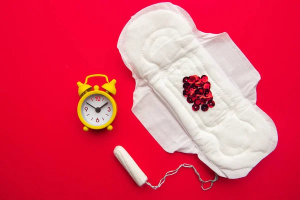 Top view of feminine pad and cotton tampon with red glitters and yellow alarm clock on red background