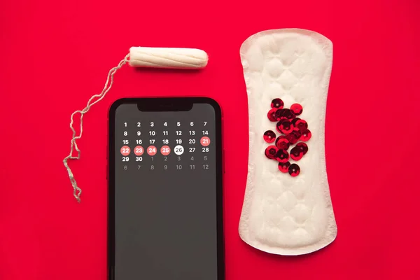 Mobil alkalmazás nyomon követni a menstruációs ciklus és a jelek. PMS és a kritikus napok koncepciója. Pamut tampon, egészségügyi betét, piros csillogás a piros háttér. — Stock Fotó
