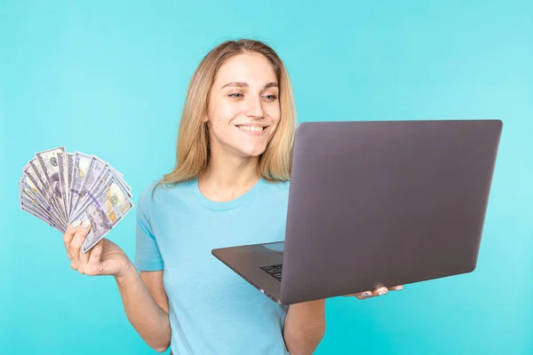 Młoda kobieta pracująca na nowoczesnym laptopie, trzymająca wiązkę dolarów, gotówkę odizolowaną na niebieskim tle. Finanse, praca na własny rachunek, mobilne biuro — Zdjęcie stockowe