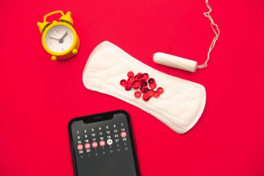 Adet döngüsü. Akıllı telefon ekranında mobil uygulama işaretleri olan bir ay takvimi. Alarm, pamuk tampon, hijyenik ped, kırmızı arka planda kırmızı parıltılar.