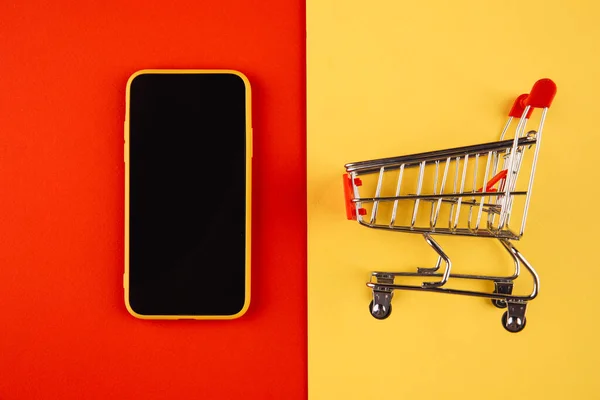 Conceptos de compras en línea con carro de maqueta y teléfono inteligente sobre fondo rojo amarillo. Mercado de comercio electrónico. Transporte logístico. Comercio minorista — Foto de Stock