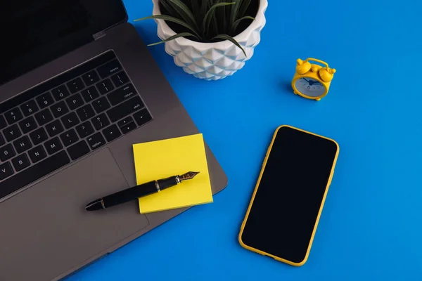 Концепция рабочего места домашнего офиса. Ноутбук, часы, смартфон, липкая нота и ручка на синем фоне — стоковое фото
