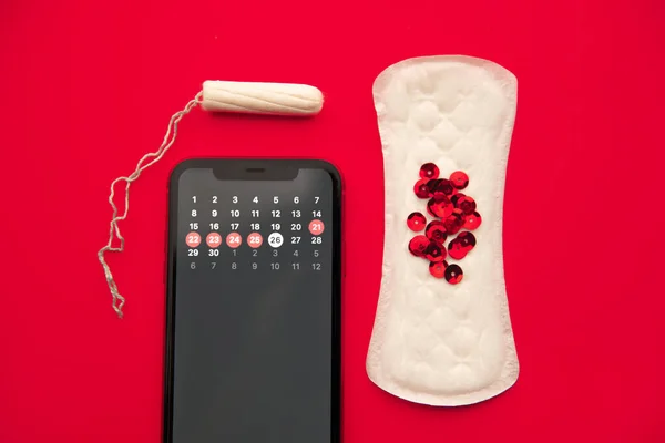 Menstruatiecyclusconcept. Kalender voor de maand met markeringen in mobiele applicatie op het smartphone scherm. Alarm, katoenen tampon, sanitairpad, rode glitters op de rode achtergrond — Stockfoto
