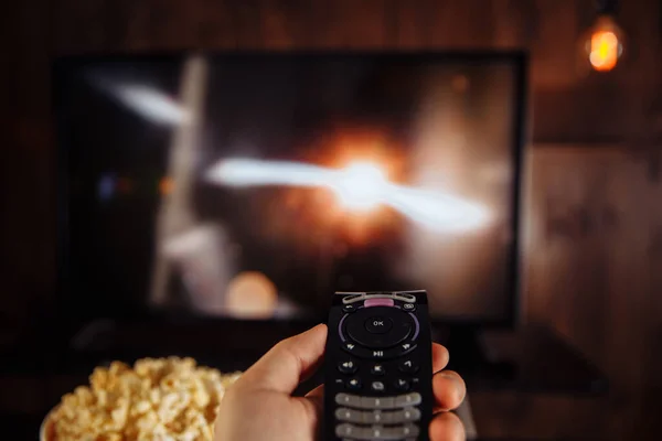 Pria dengan TV remote control duduk di rumah dengan semangkuk besar popcorn. — Stok Foto