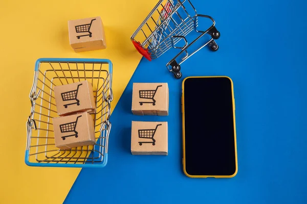 Conceito de compras online. Smartphone com cinco mini-caixas, cesta de compras e carrinho em fundo amarelo azul — Fotografia de Stock