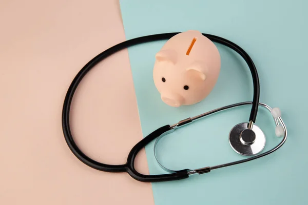 Medicine doctor equipment stethoscope and piggy bank isolated on pink blue pastel background. Health care financial checkup or saving for medical insurance costs concept