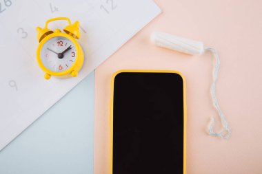 Adet döngüsü konsepti. Sarı alarm saati ve akıllı telefon ekranında mobil uygulama. Arka planda pamuk tampon